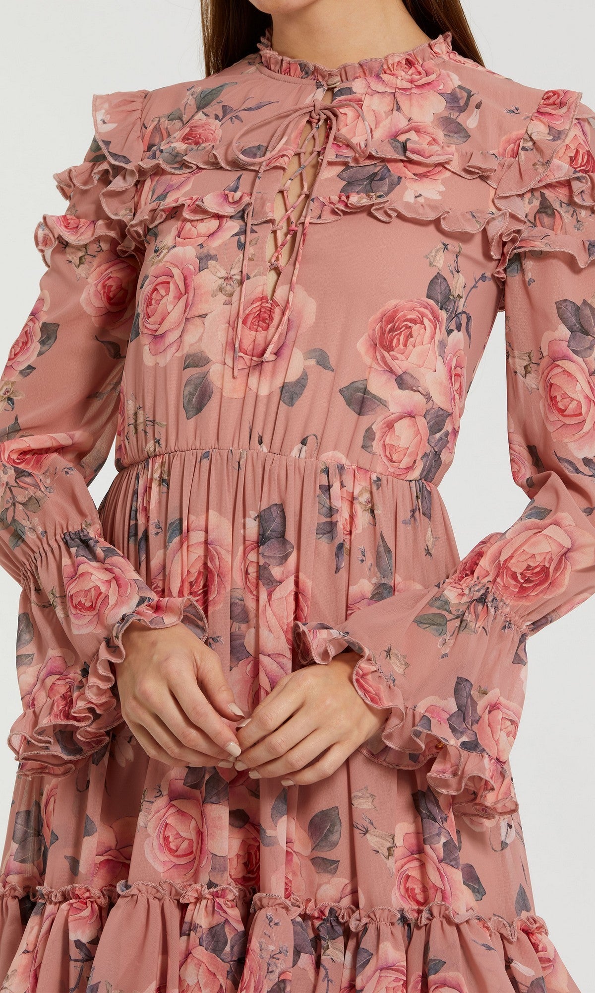 A person wearing the Mac Duggal 9306 Midi Wedding Guest Dress, featuring long sleeves, ruffles, a pink floral chiffon fabric, and a lace-up neckline. Their hands are folded at the waist and only the upper body is visible.